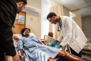 There is a patient laying in a hospital bed and two students are examining the patient
