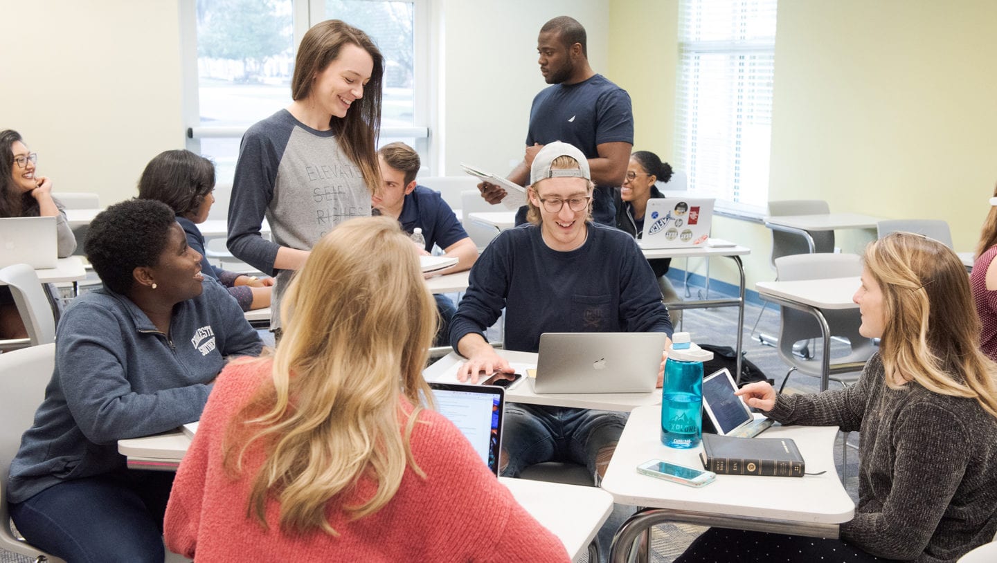 Students standing and sitting at desks circled in a group and talking with each other.