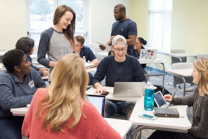 Students standing and sitting at desks circled in a group and talking with each other.