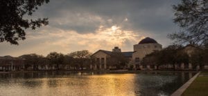 The Science Building with the sun setting behind it casting rays through the clouds. The golden color reflects off of the reflection pond.