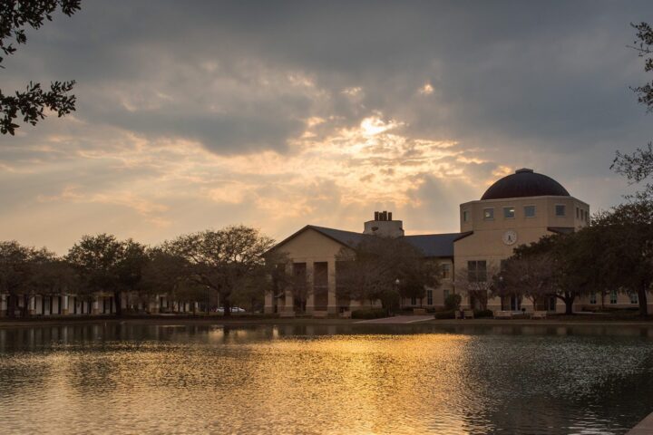 The Science Building with the sun setting behind it casting rays through the clouds. The golden color reflects off of the reflection pond.