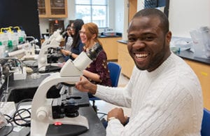 Students smiling in a biology lab classroom as the sit in front of microscopes.