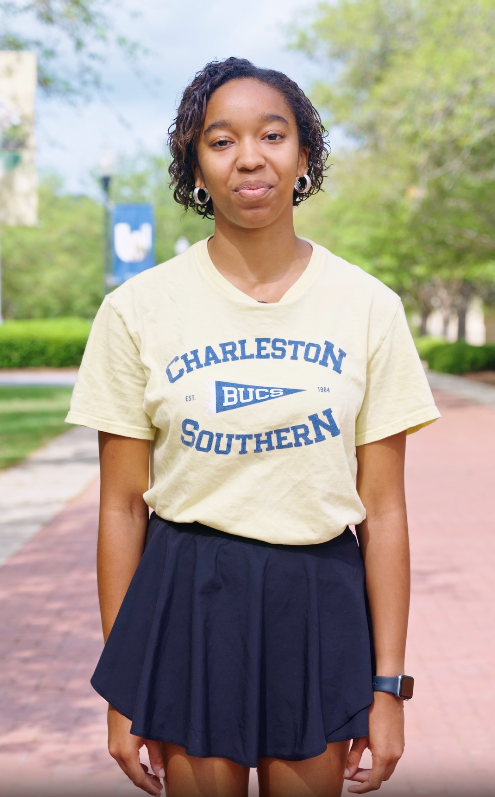 Black female student with short hair and a yellow t-shirt stands on a brick walkway facing the camera