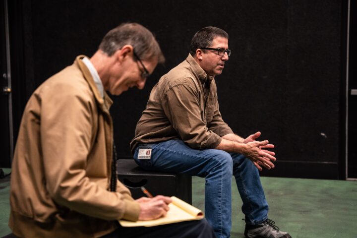 A professor sitting on a box explainig the play called the Holocaust and a man taking notes
